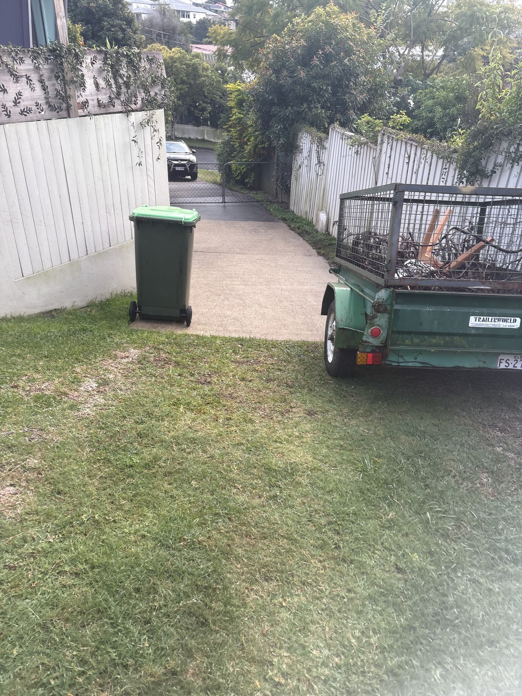 Side laneway driveway leading past a wheelie bin to a loaded garden-waste trailer.